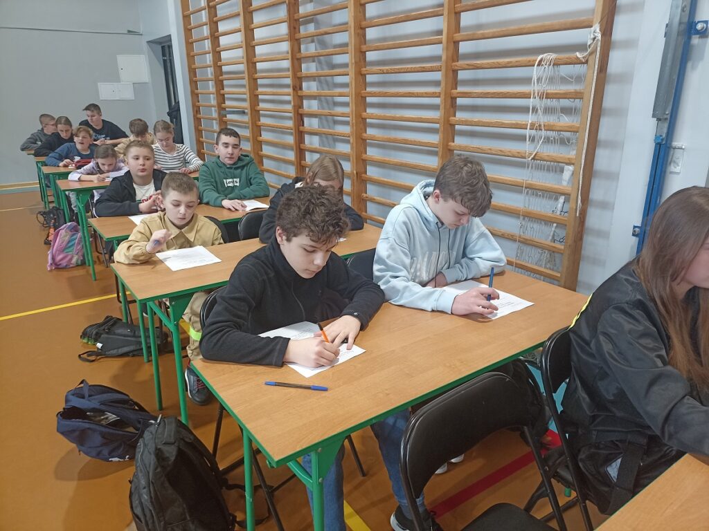 Uczniowie piszący test na sali gimnastycznej podczas eliminacji gminnych XLVII Ogólnopolskiego Turnieju Wiedzy Pożarniczej "Młodzież zapobiega pożarom"