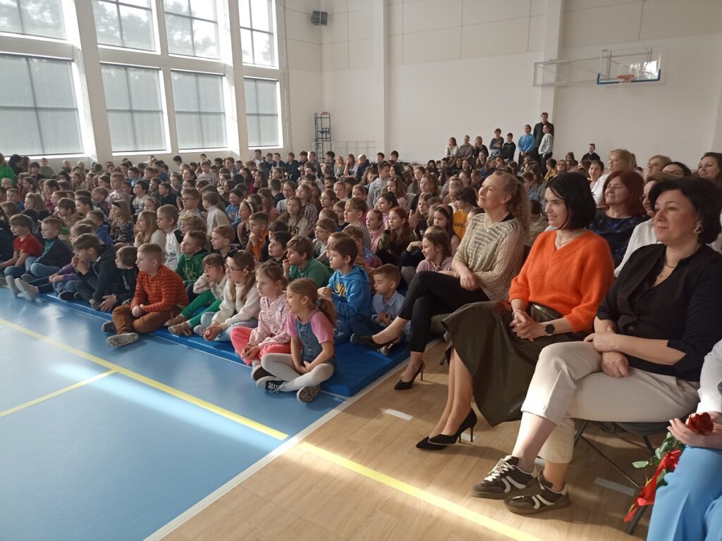 Dyrekcja Szkoły z gronem pedagogicznym w w PSP nr 3 z O. I. w Kozienicach. Obok na materacach siedzący uczniowie, którzy patrzą w kierunku występujących dzieci na akademii z okazji Dnia Kobiet