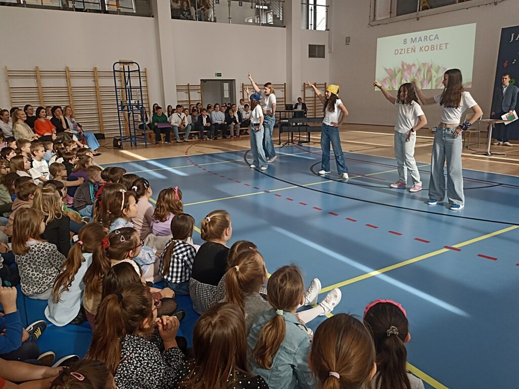 Pięć dziewcząt, które tańczą w rytm piosenki podczas akademii z okazji Dnia Kobiet w PSP nr 3 z O. I. w Kozienicach. W tle dyrekcja szkoły z gronem pedagogicznym i oglądającymi uczniami