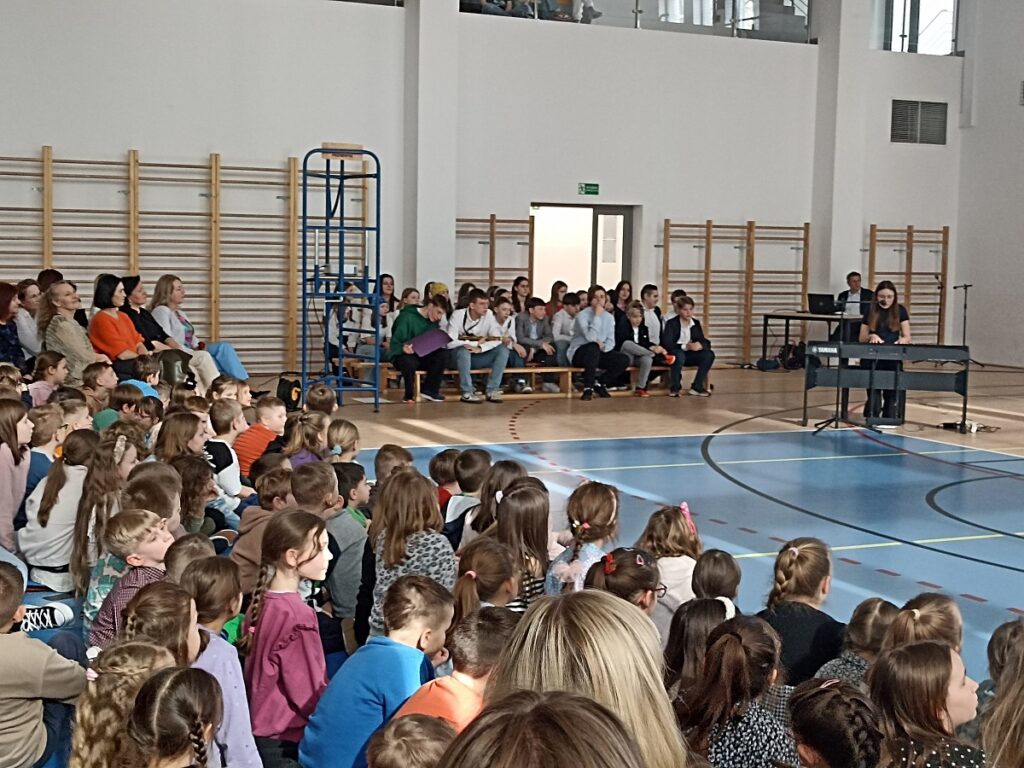 Występujący uczennica grająca na instrumencie klawiszowym (keyboard) na akademii z okazji Dnia Kobiet w PSP nr 3 z O. I. w Kozienicach. W tle grono pedagogiczne i inni uczniowie.