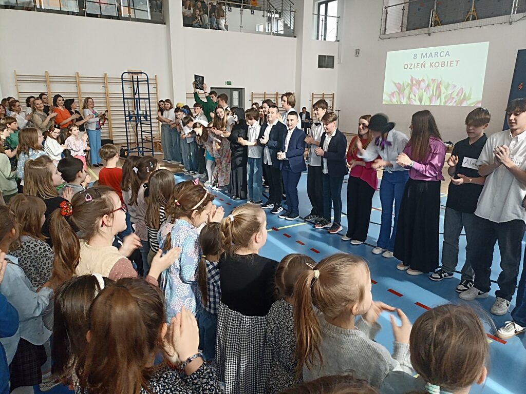 Ostatnia scena z akademii z okazji Dnia Kobiet w PSP nr 3 z O. I. w Kozienicach. W tle inni uczniowie. Kilkanaście występujących osób zadowolonych ze swoich występów. W tle grono pedagogiczne i i inni uczniowie oklaskujący występujących