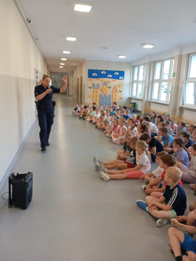 Zdjęcie przedstawia dzieci, nauczycieli oraz panią policjant na korytarzu szkolnym.