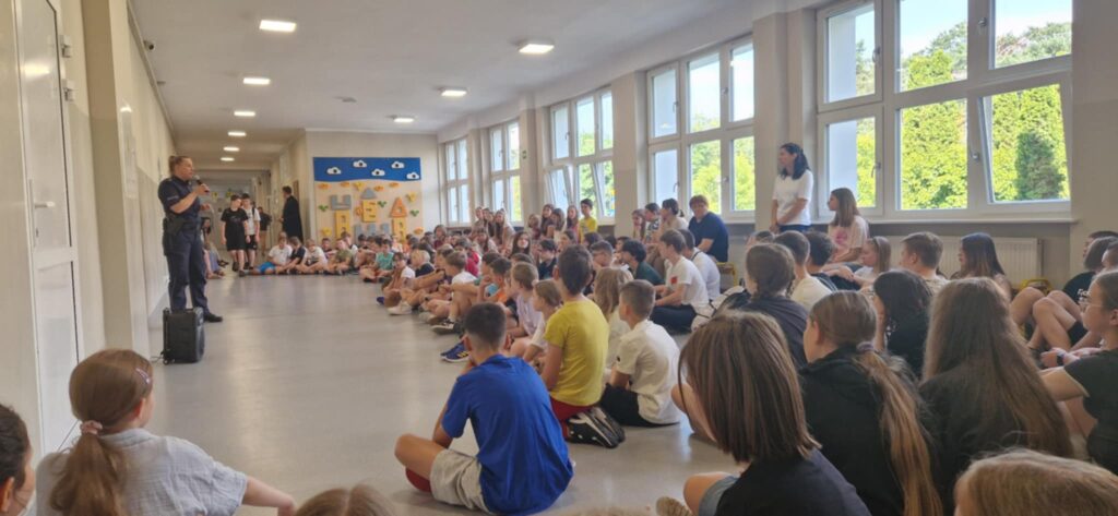 Zdjęcie przedstawia dzieci, nauczycieli oraz panią policjant na korytarzu szkolnym.